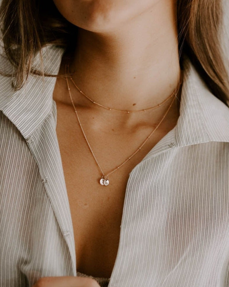A woman wears a Rose Gold Petite Personalised Necklace layered with a 35cm Rose Gold Ball Chain Necklace for an effortless personalised layered necklace look with an open collared light-weight shirt.