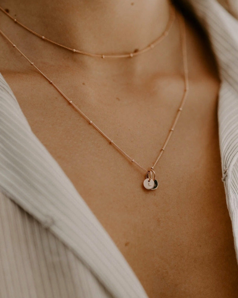 A close up side profile of the Rose Gold Petite Personalised necklace sitting elegantly on a woman's décolletage. The necklace is woern at a 45cm mid length with a shorter, 35cm length, Rose Gold Ball Chain necklace layered with it.