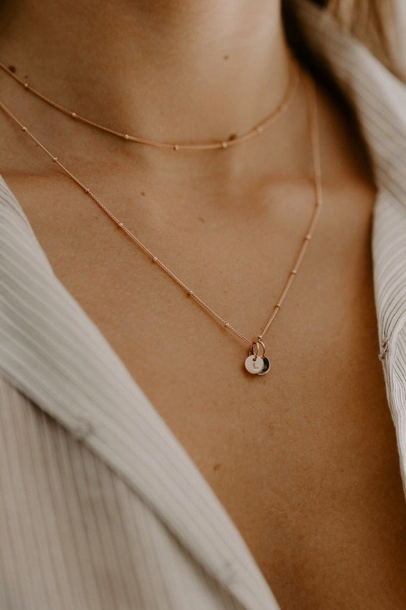 A close up side profile of the Rose Gold Petite Personalised necklace sitting elegantly on a woman's décolletage.  The necklace is woern at a 45cm mid length with a shorter, 35cm length, Rose Gold Ball Chain necklace layered with it.