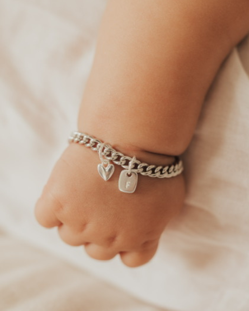 The sweetest close up of a baby wearing a Sweetheart Cuban Link bracelet that has a mini sweetheart pendant on it as well as a mini rectangular initial pendant by Bluebird Jewellery