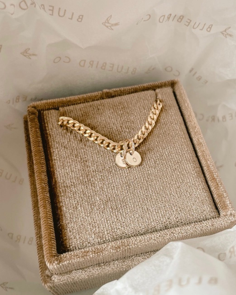 A gold bracelet with personalised pendants, placed inside a Bluebird Co. brown velvet box by Bluebird Jewellery