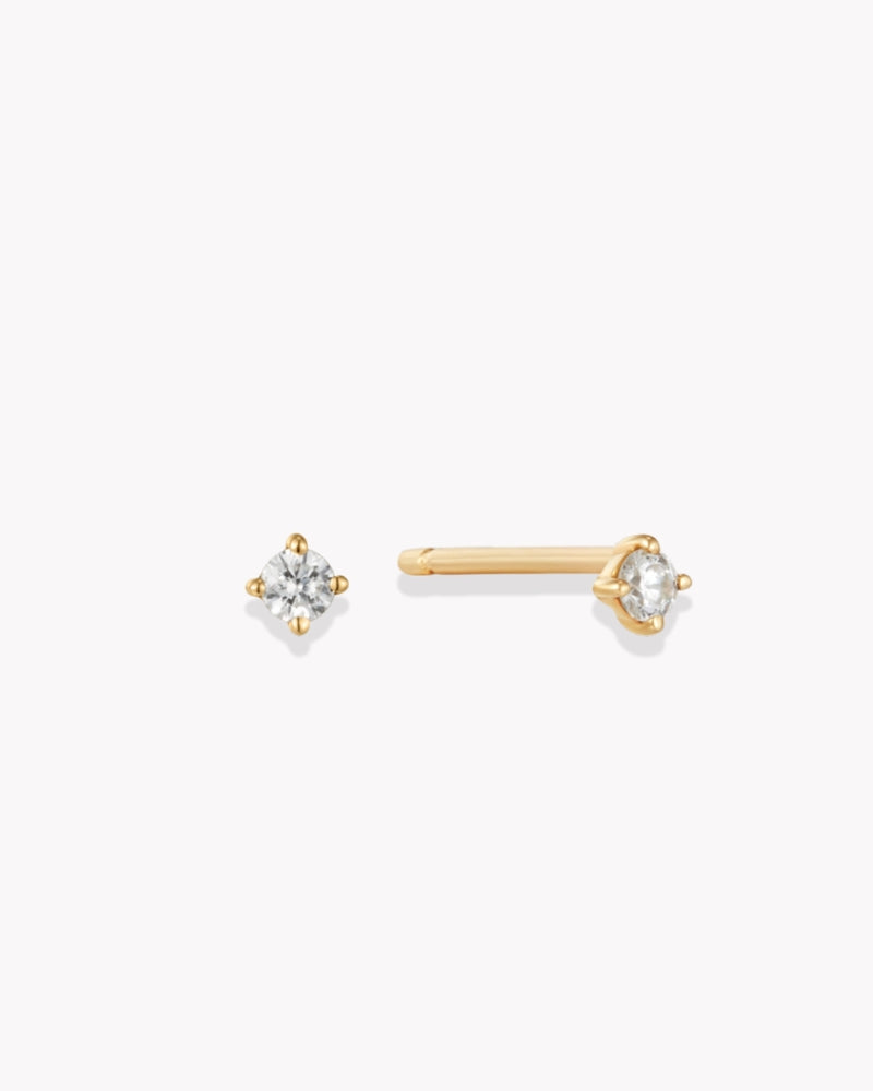 A pair of dainty gold stud natural diamond earrings with a shimmering diamond in a classic four-prong setting, captured on a white background.