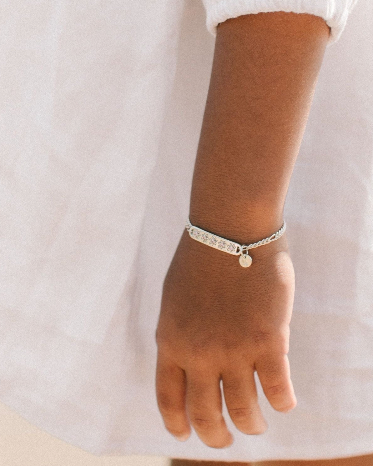 A child wearing a silver Daisy Bracelet by Bluebird Jewellery, featuring five hand-drawn daisies with pink cubic zirconia centres, crafted as a feminine and floral fine jewellery piece along with an initial pendant.