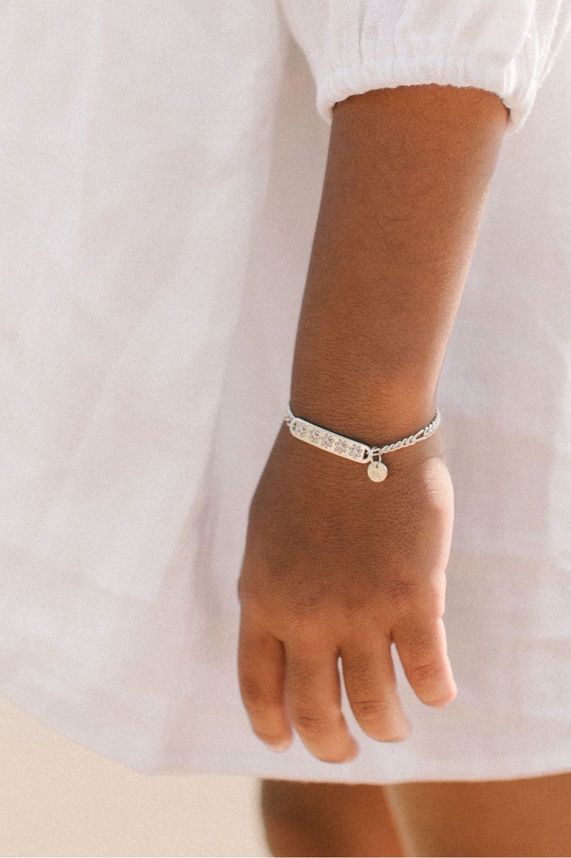 A child wearing a silver Daisy Bracelet by Bluebird Jewellery, featuring five hand-drawn daisies with pink cubic zirconia centres, crafted as a feminine and floral fine jewellery piece along with an initial pendant.