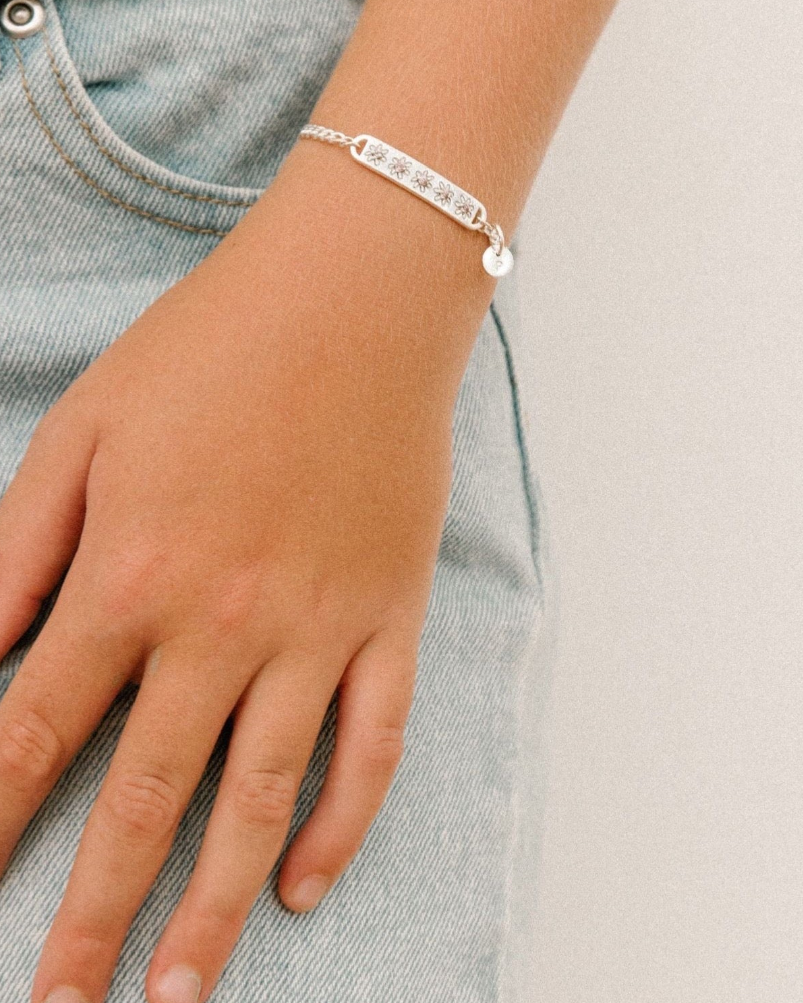 A child wearing a silver Daisy Bracelet by Bluebird Jewellery, featuring five hand-drawn daisies with pink cubic zirconia centres, crafted as a feminine and floral fine jewellery piece along with an initial pendant that represents her name.