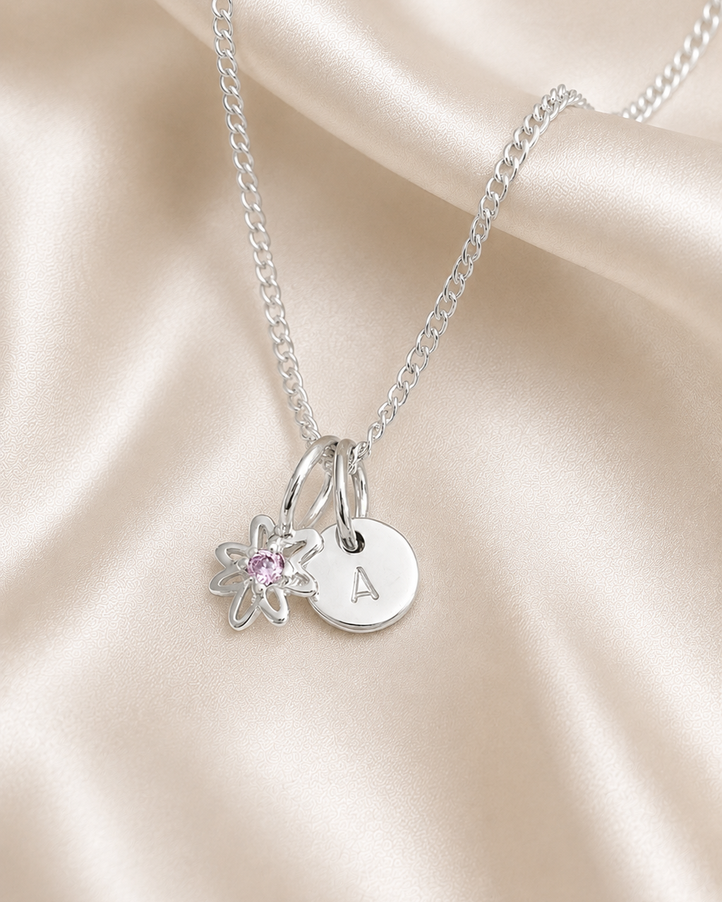 A silver chain necklace with two charms: a round engraved disc stamped with the letter 'A' and a small flower charm with a pink gemstone, displayed on a draped beige satin fabric.