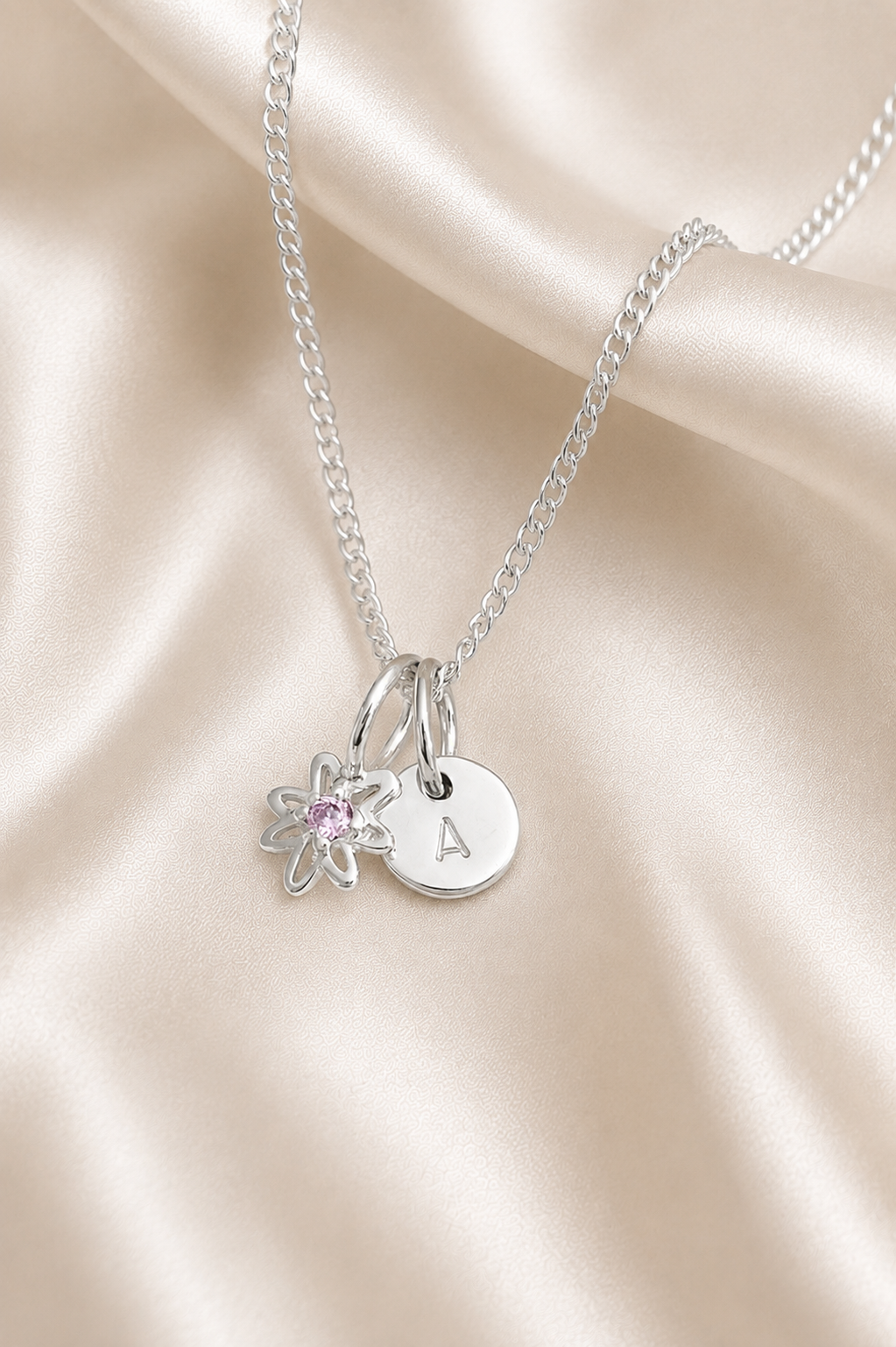 A silver chain necklace with two charms: a round engraved disc stamped with the letter 'A' and a small flower charm with a pink gemstone, displayed on a draped beige satin fabric.