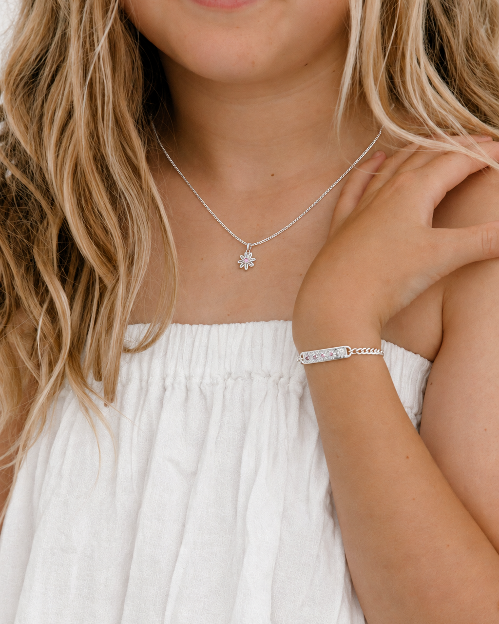 A close-up lifestyle shot of a young girl with blonde hair wearing a delicate silver necklace with a small flower-like pendant and a matching silver bracelet.
