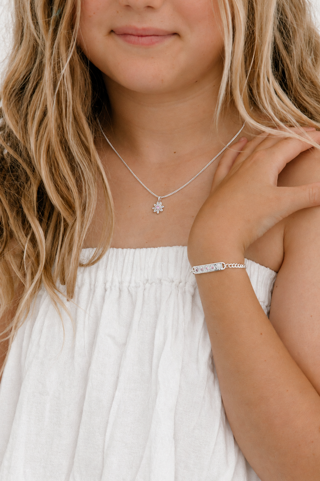 A close-up lifestyle shot of a young girl with blonde hair wearing a delicate silver necklace with a small flower-like pendant and a matching silver bracelet.