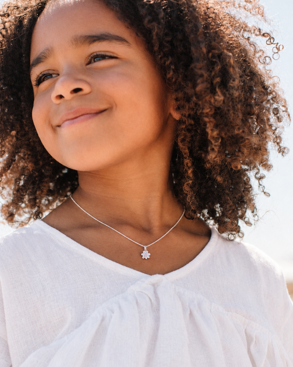 A close-up lifestyle shot of a young girl with curly brown hair wearing a delicate silver necklace with a small flower-shaped pendant, paired with a white top.