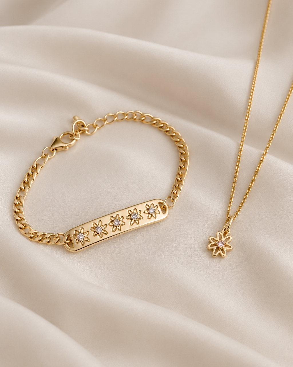 Gold jewelry set featuring a bar bracelet with five star-shaped crystal accents and a delicate chain necklace with a small flower pendant, displayed on draped cream satin fabric.