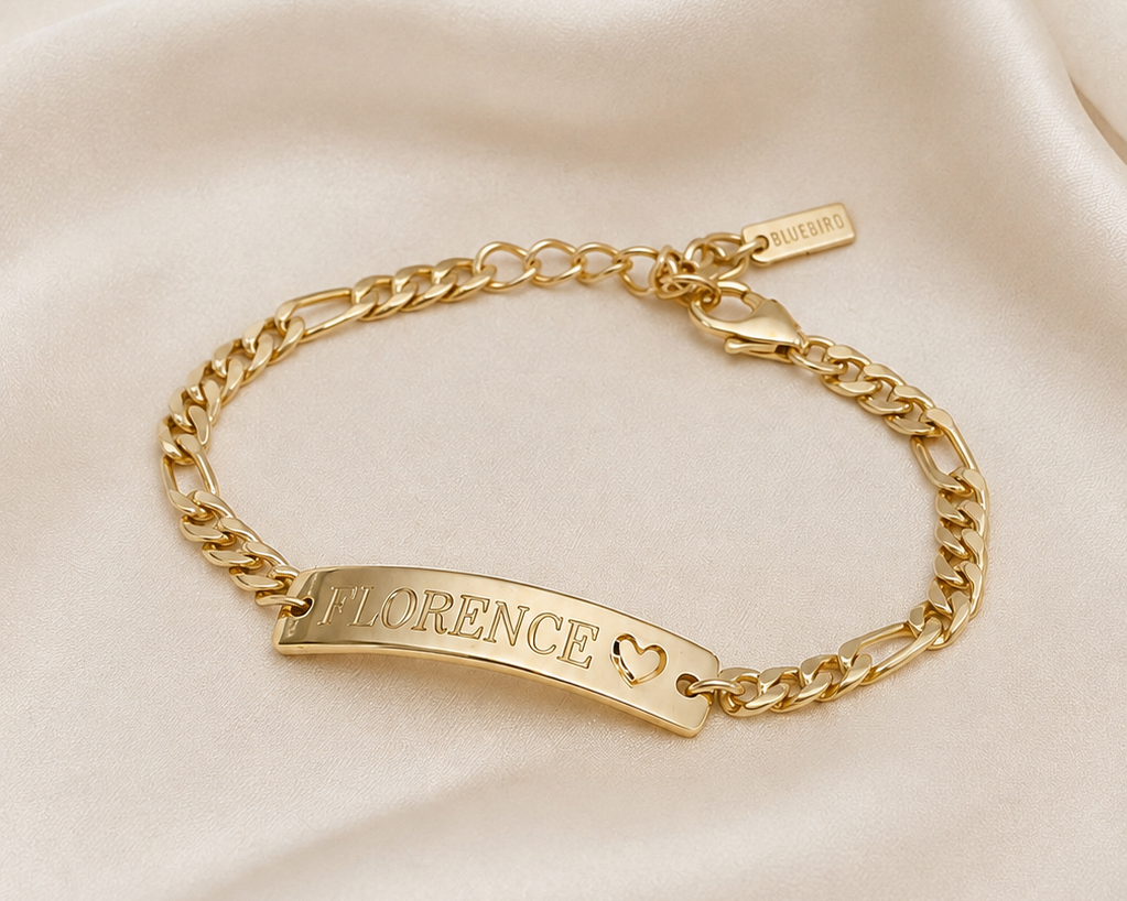 A gold chain bracelet with a curved ID plate engraved with the name 'FLORENCE' and a small heart, displayed on a draped cream satin fabric background.