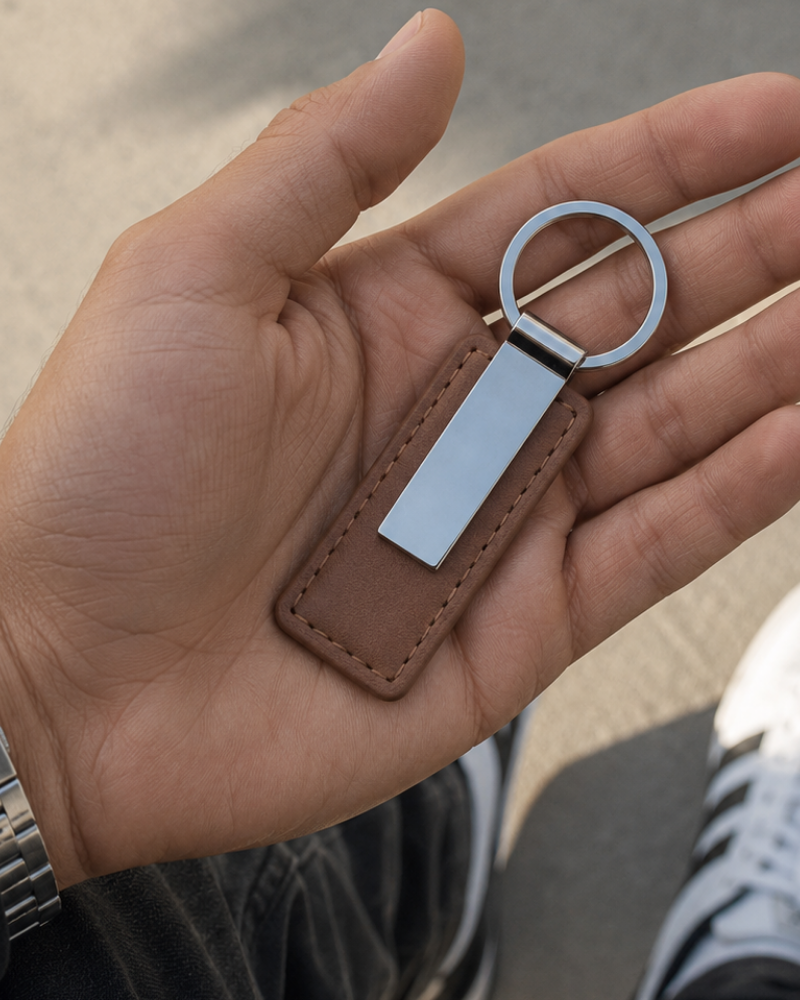 A close-up of a hand holding a blank keychain with a brown leather tag and a silver metal key ring and plate, set against an outdoor pavement background.