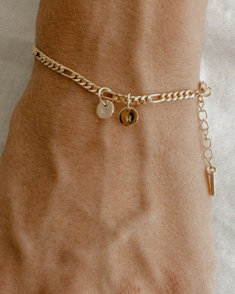 A Figaro Bracelet with two solid gold 6mm initial pendants rests elegantly on a woman's wrist by Bluebird Jewellery