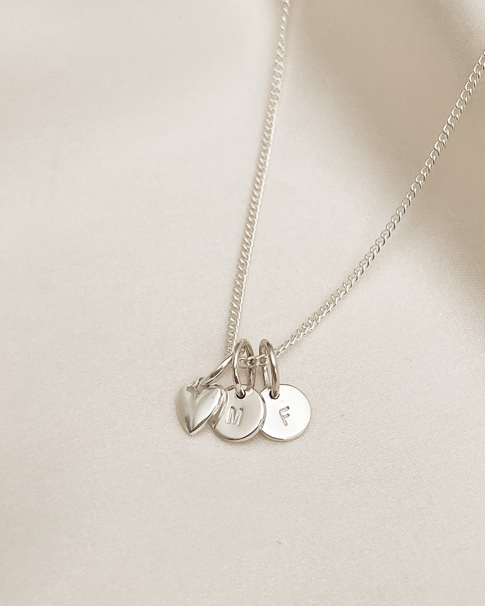 A silver fine curb chain has a mini sweetheart pendant and two initial pendants on it.