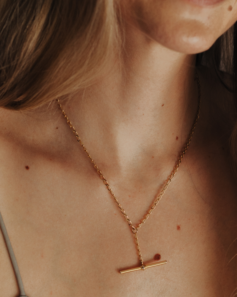 A Gold Fob Necklace also known as a T-Bar Necklace is worn on a woman and features an elegant curb chain with T-Bar drop chain moving freely along the chain.