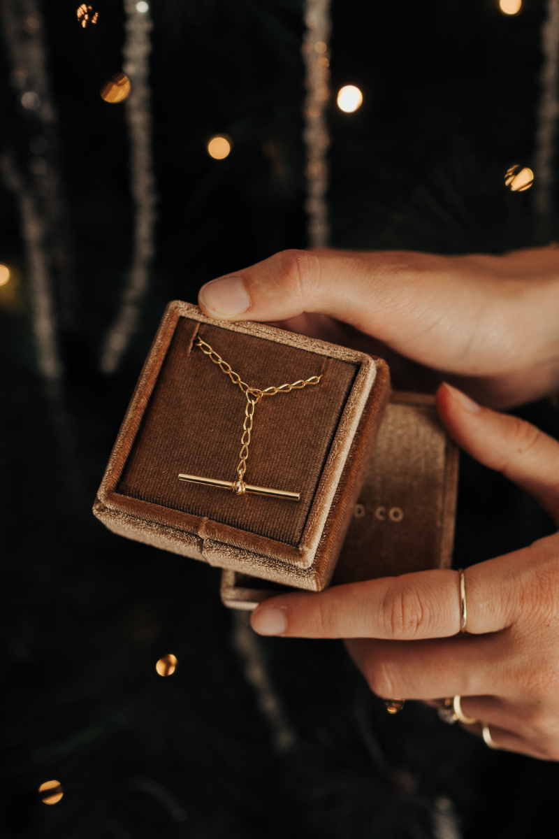 A gold Fob necklace is resting inside a Bluebird Co velvet gift box and a woman's hands are holding the gift box.