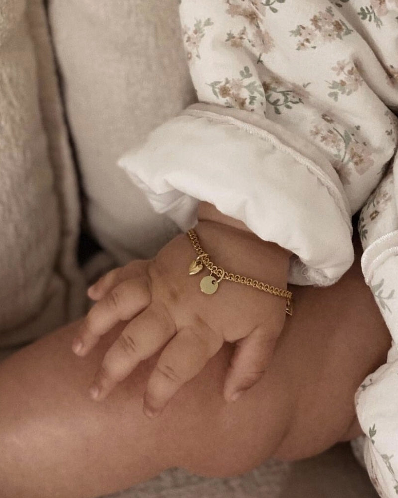 A close-up of a baby’s wrist wearing a Lace Chain Bracelet with a heart charm and round initial pendant, crafted as a delicate personalised keepsake by Bluebird Jewellery