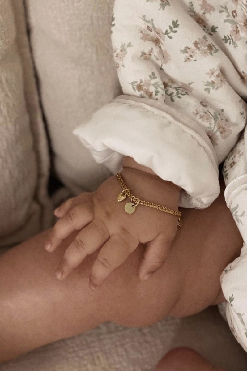 A close-up of a baby’s wrist wearing a Lace Chain Bracelet with a heart charm and round initial pendant, crafted as a delicate personalised keepsake.