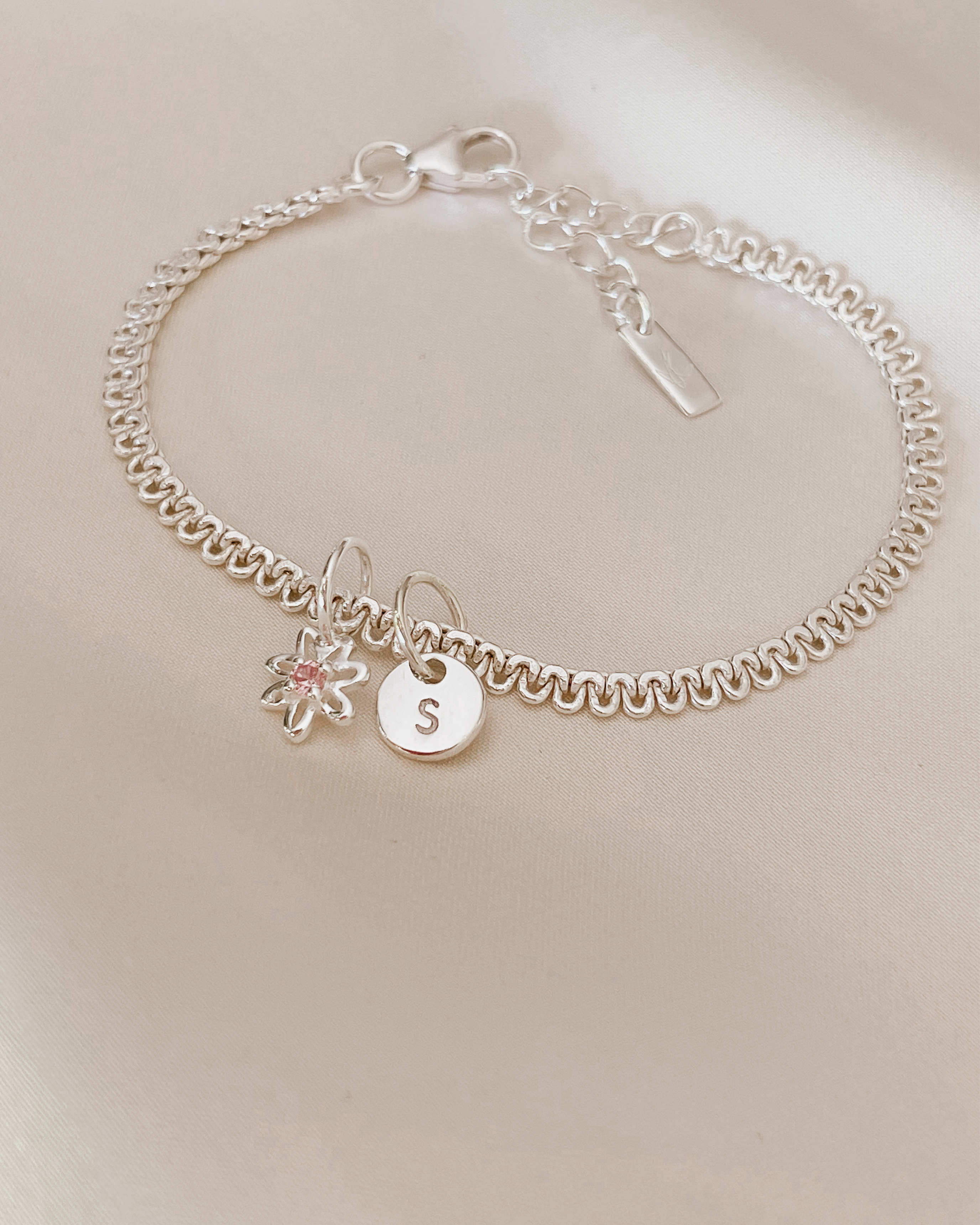 A lace chain bracelet is shown ip close on silk with a beautiful Daisy Pendant taht has a pink cubic zirconia on it and a Petite Initial Pendant by Bluebird Jewellery