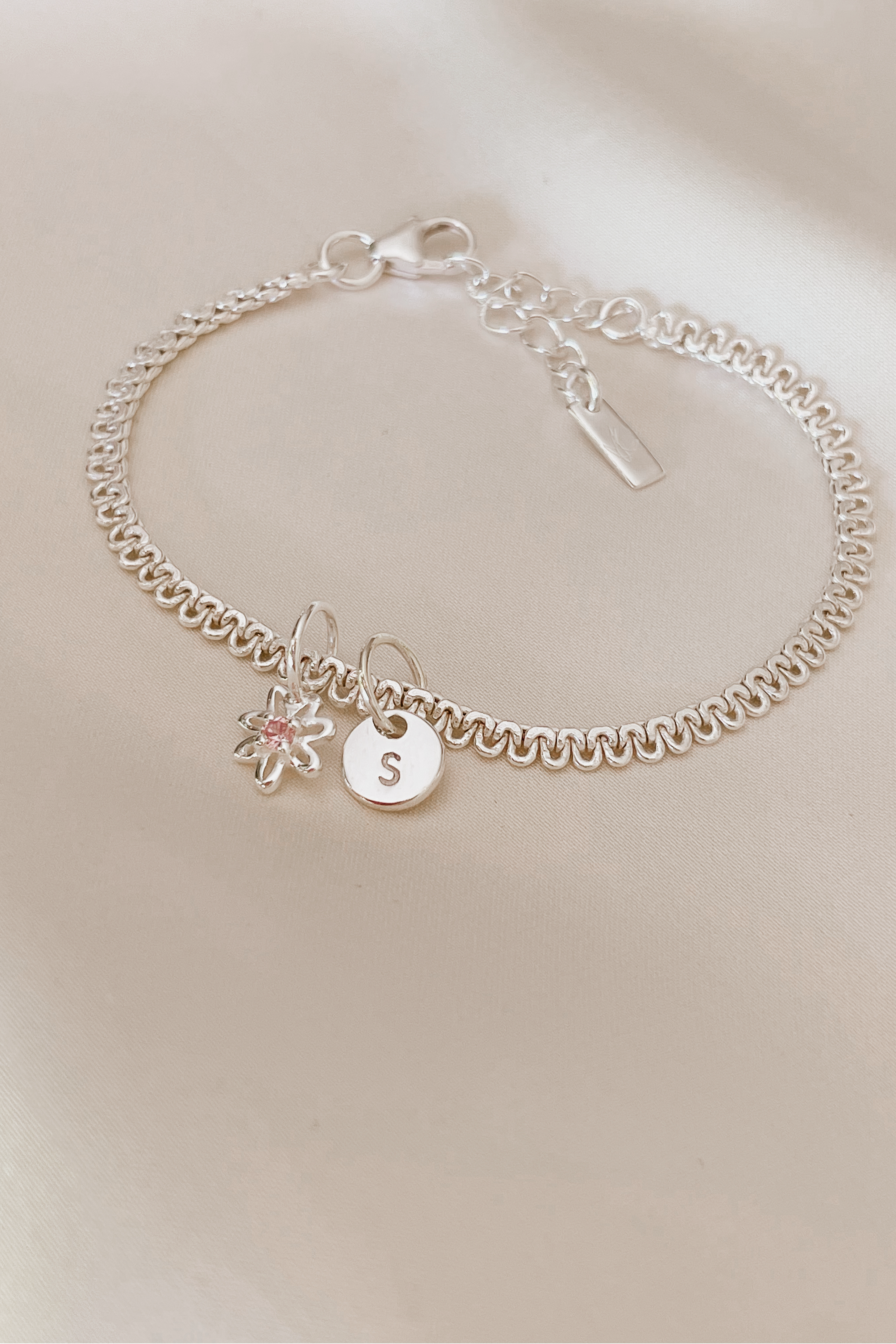 A lace chain bracelet is shown ip close on silk with a beautiful Daisy Pendant taht has a pink cubic zirconia on it and a Petite Initial Pendant by Bluebird Jewellery