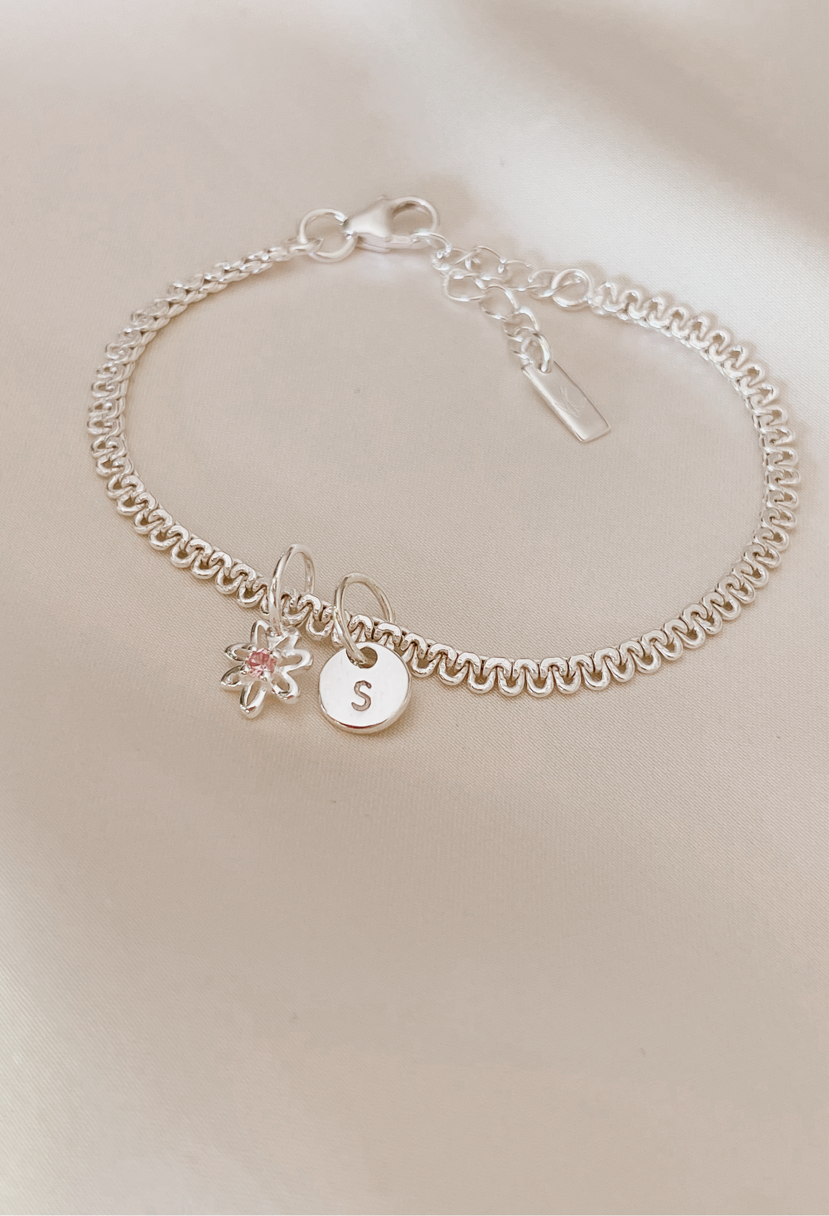 A lace chain bracelet is shown ip close on silk with a beautiful Daisy Pendant taht has a pink cubic zirconia on it and a Petite Initial Pendant.