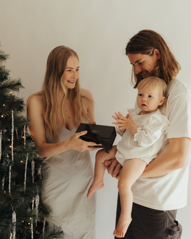 Baby is wearing a gold anklet and is being held by her dad as her family stands at the Christmas tree. The anklet has a lace chain style.