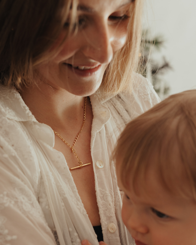 A woman holding her child is wearing a Fob Necklace in Gold