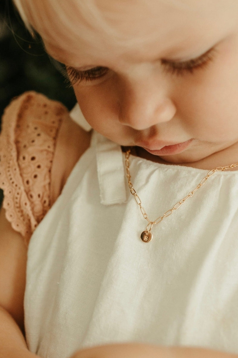 A baby wearing a link chain necklace with a classic round Petite Gold Vermeil Initial Pendant from Bluebird Jewellery, personalised with your chosen letter and designed to be worn elegantly on any necklace, bracelet, or anklet chain.