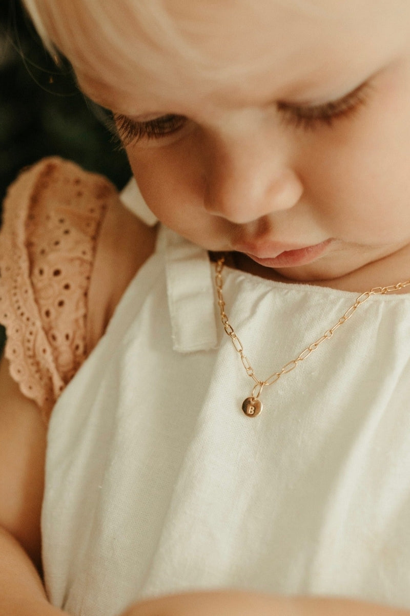A baby wearing a link chain necklace with a classic round Petite Gold Vermeil Initial Pendant from Bluebird Jewellery, personalised with your chosen letter and designed to be worn elegantly on any necklace, bracelet, or anklet chain.
