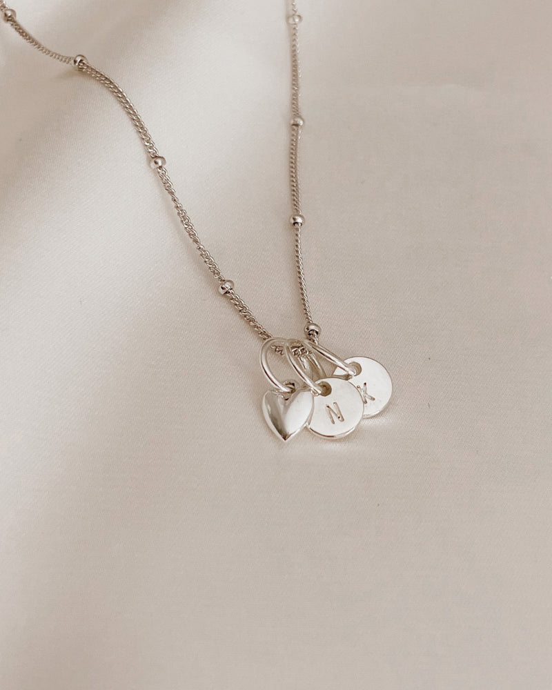 A sterling silver necklace chain with a heart pendant and two petite initial pendants that are round.