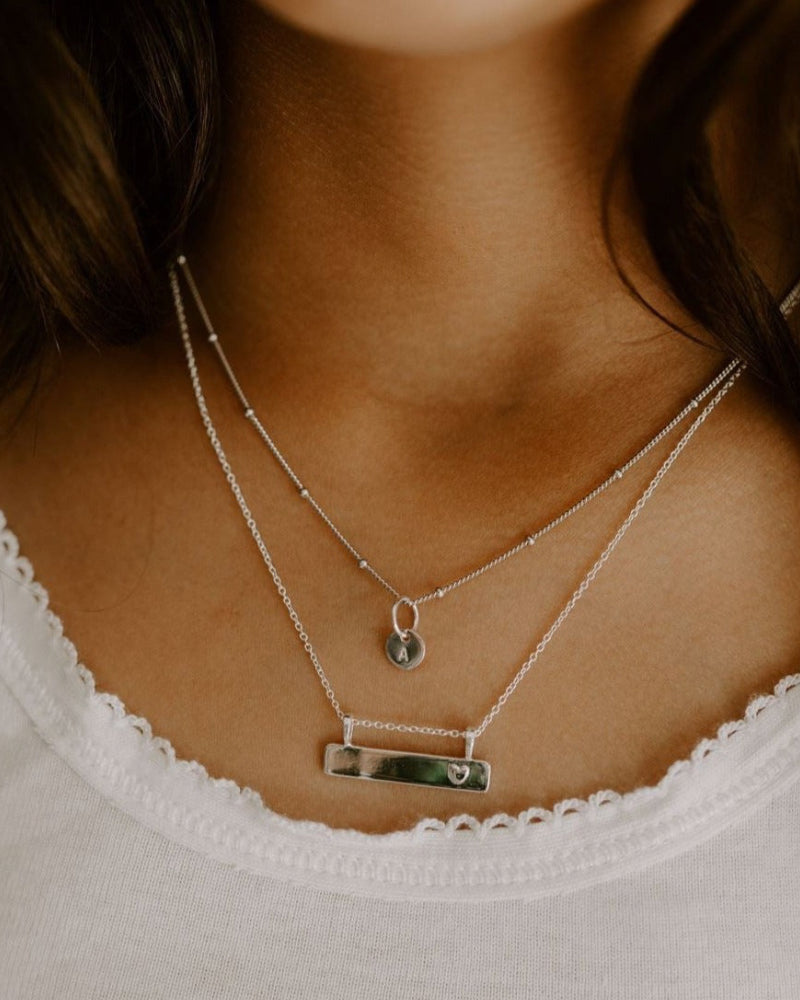 A child wears the petite personalised necklace with one initial pendant. The necklace is layered with the Darling Bar Necklace also in sterling silver. Both necklaces can be customised with personalised detailing.