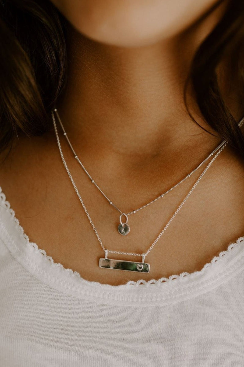 A child wears the petite personalised necklace with one initial pendant. The necklace is layered with the Darling Bar Necklace also in sterling silver. Both necklaces can be customised with personalised detailing.