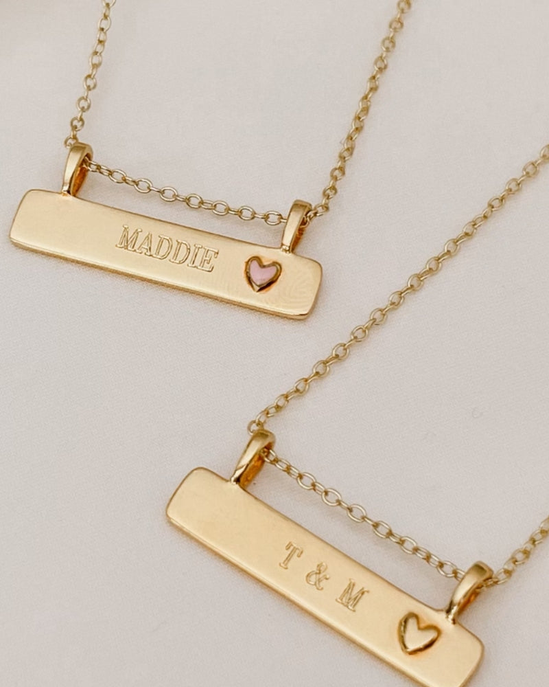 A Pink Darling Bar Necklace sits alongside a Darling Bar Necklace. Both necklaces have personalised engraving on the front of them.