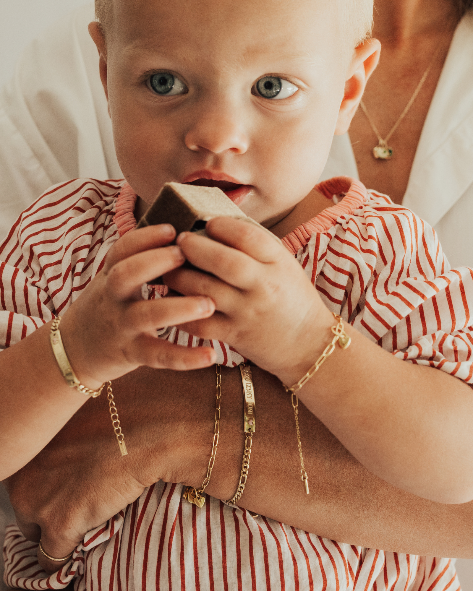 A tender moment featuring a baby and mum wearing matching gold bracelets — the Pink Darling and Posie — with heart charms, as the baby holds a velvet box by Bluebird Jewellery.