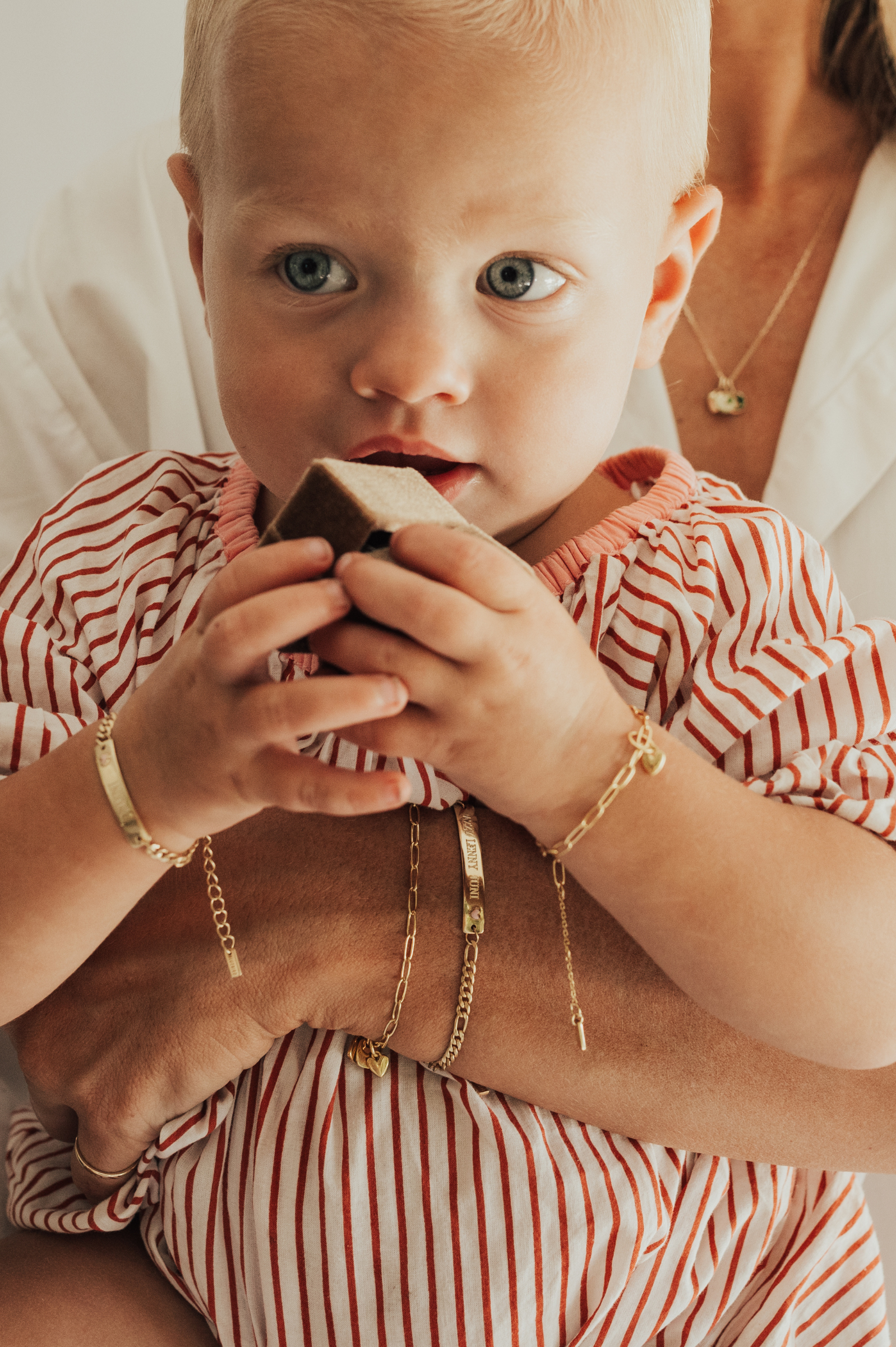 A tender moment featuring a baby and mum wearing matching gold bracelets — the Pink Darling and Posie — with heart charms, as the baby holds a velvet box.