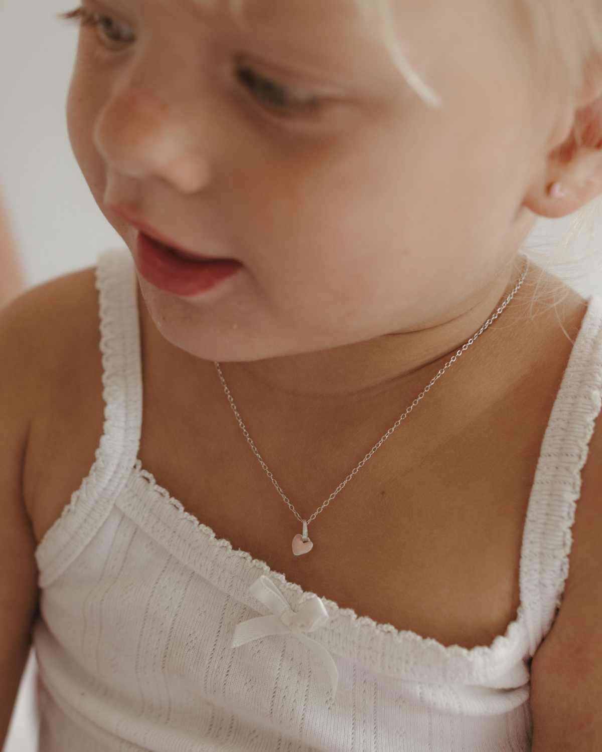 A young girl wears a soft pink coloured heart pendant necklace on a solid sterling silver chain. It can be personalised with an initial pendant