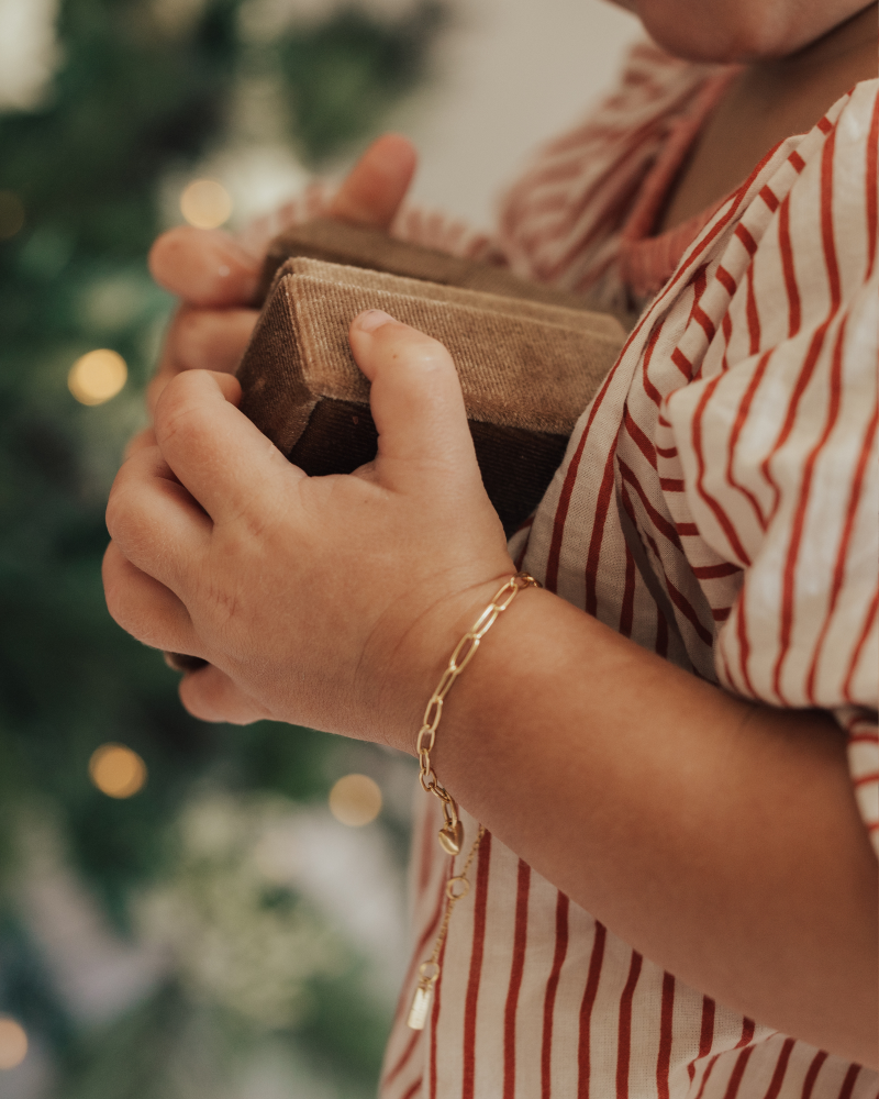 A baby wearing a gold Posie Bracelet with a Mini Sweetheart pendant, holding a velvet box captured in natural light to highlight its delicate craftsmanship by Bluebird Jewellery.