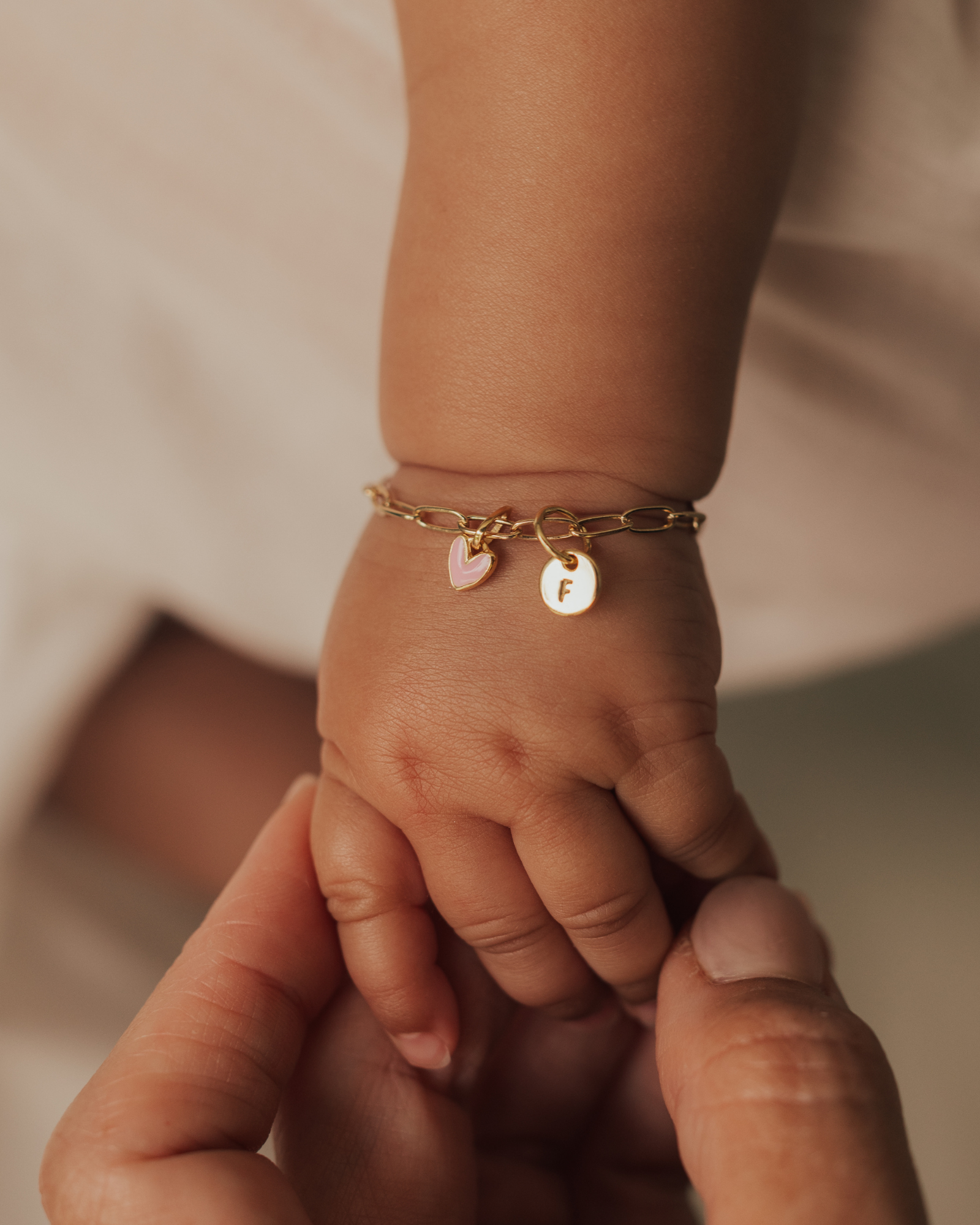 A mother is holding a baby's hand and the baby is wearing a gold baby bracelet with pink heart pendant and petite round initial pendant. Bluebird Jewellery.