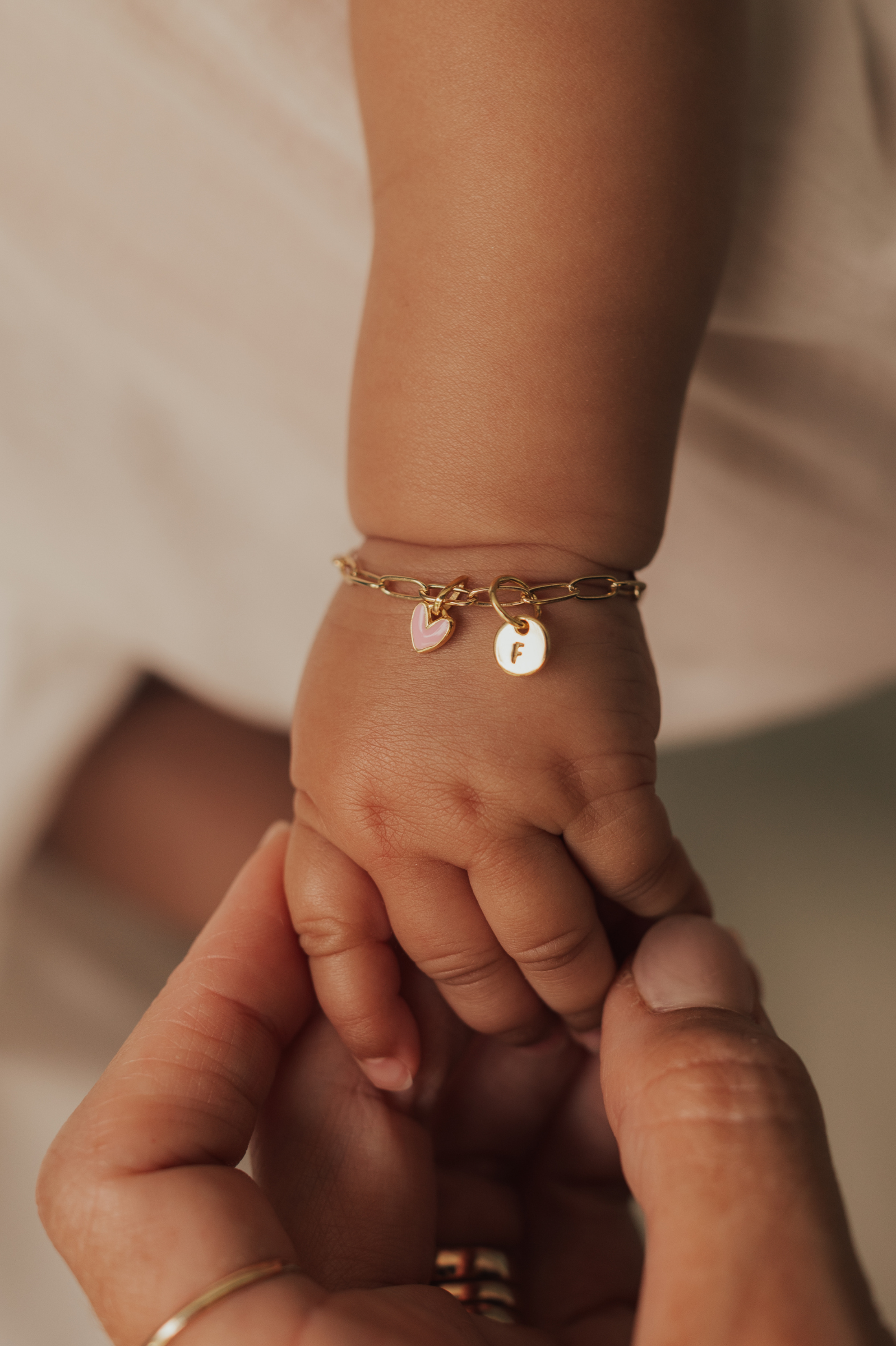 A mother is holding a baby's hand and the baby is wearing a gold baby bracelet with pink heart pendant and petite round initial pendant. Bluebird Jewellery.