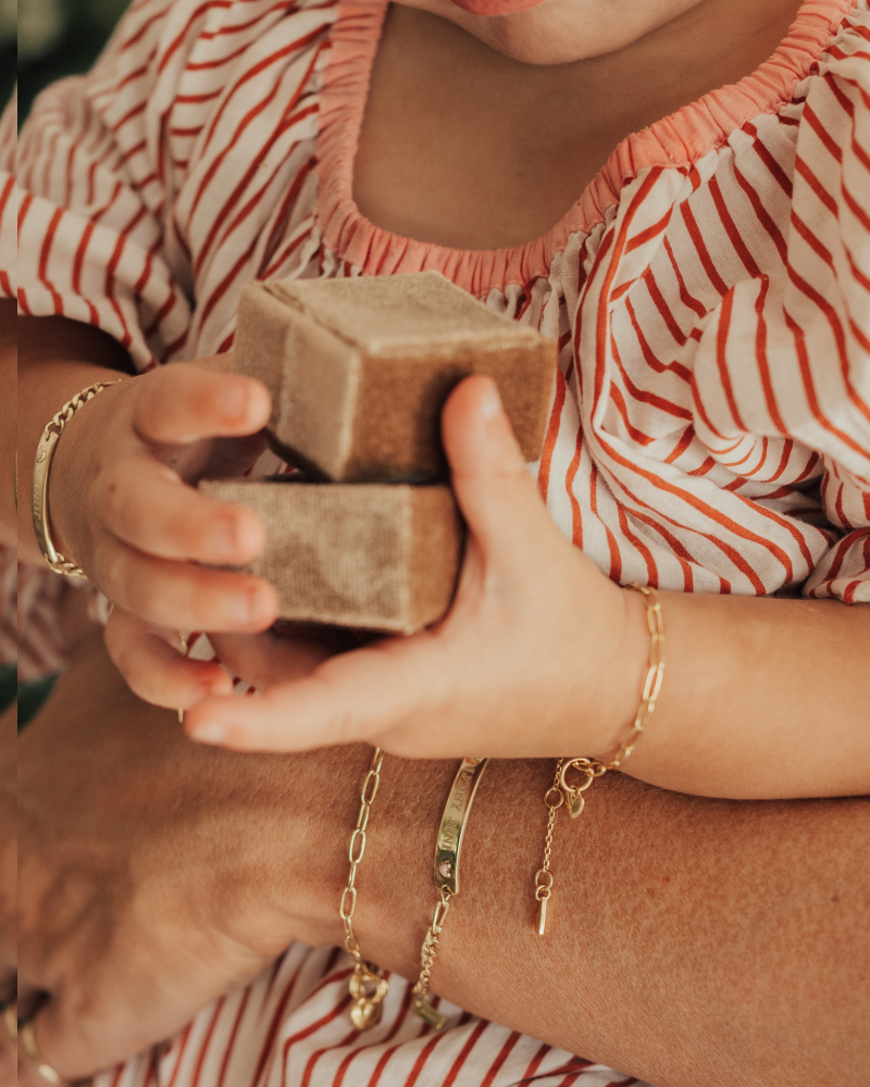 A close-up of a mother and child’s hands featuring gold link chain and personalised bar pendant bracelets, symbolising connection and timeless craftsmanship by Bluebird Jewellery.