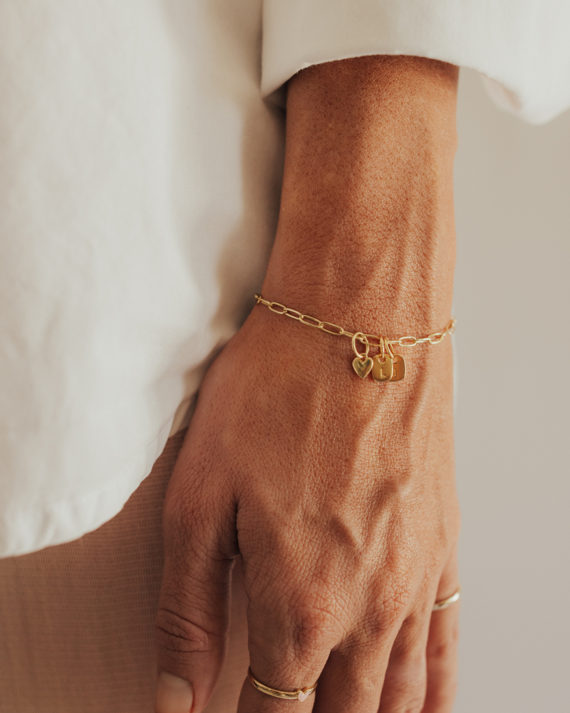 A woman wearing a gold Posie Bracelet with heart charm and engraved rectangular pendants, perfectly paired with a Darling Ring for a timeless look by Bluebird Jewellery.