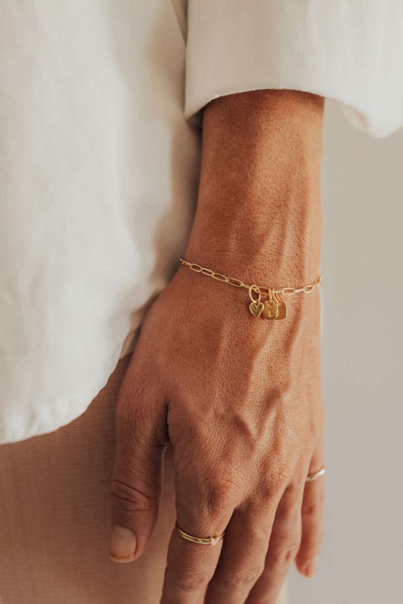 A woman wearing a gold Posie Bracelet with heart charm and engraved rectangular pendants, perfectly paired with a Darling Ring for a timeless look.