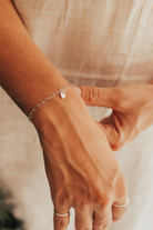 A close-up of a hand wearing a Posie Bracelet with a heart charm, complemented by delicate silver rings for a refined modern finish.