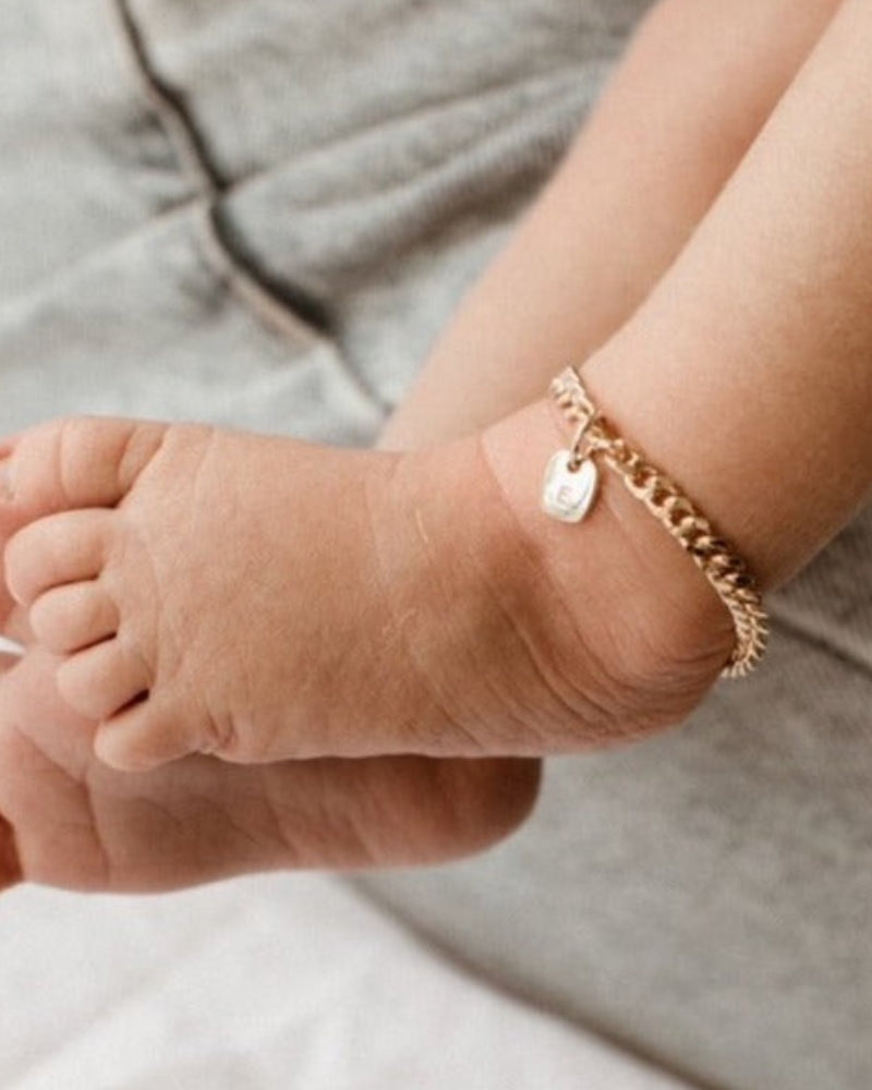 A close-up of a baby’s ankle wearing a gold Cuban Link Chain Bracelet with a solid rectangular pendant, crafted as a delicate keepsake piece.