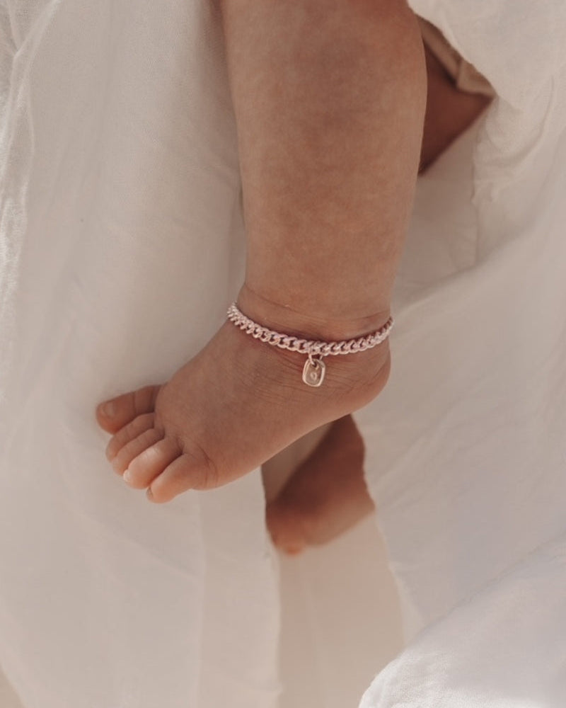 A close-up of a baby’s ankle wearing a rose gold Cuban Link Chain Bracelet with a solid gold rectangular pendant, crafted as a delicate keepsake piece.