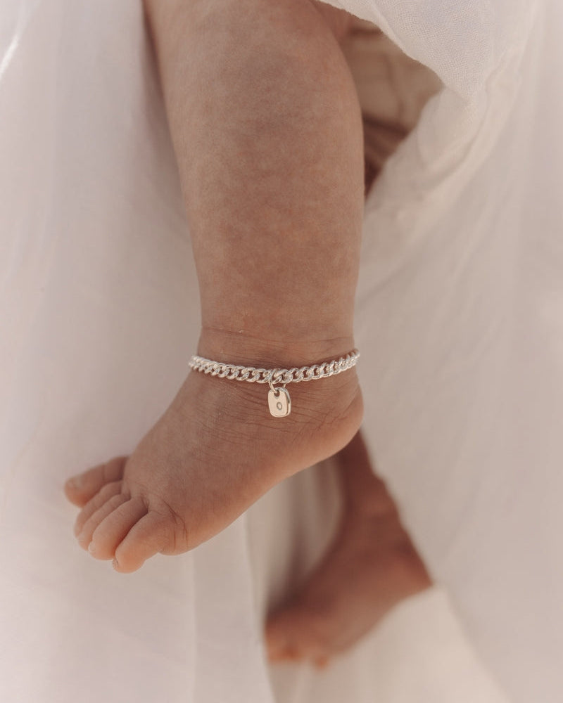 A close-up of a baby’s ankle wearing a silver Cuban Link Chain Bracelet with a rectangular pendant, crafted as a delicate keepsake piece.