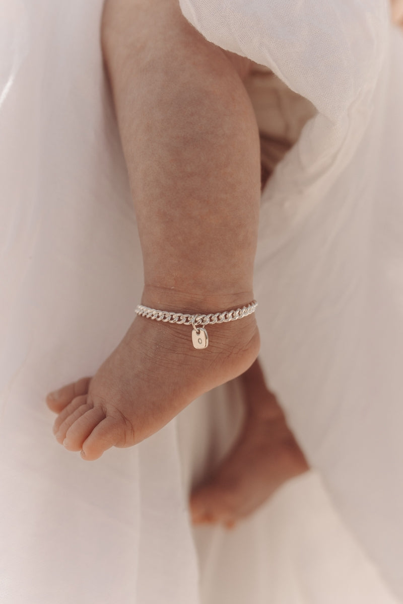 A close-up of a baby’s ankle wearing a silver Cuban Link Chain Bracelet with a rectangular pendant, crafted as a delicate keepsake piece.