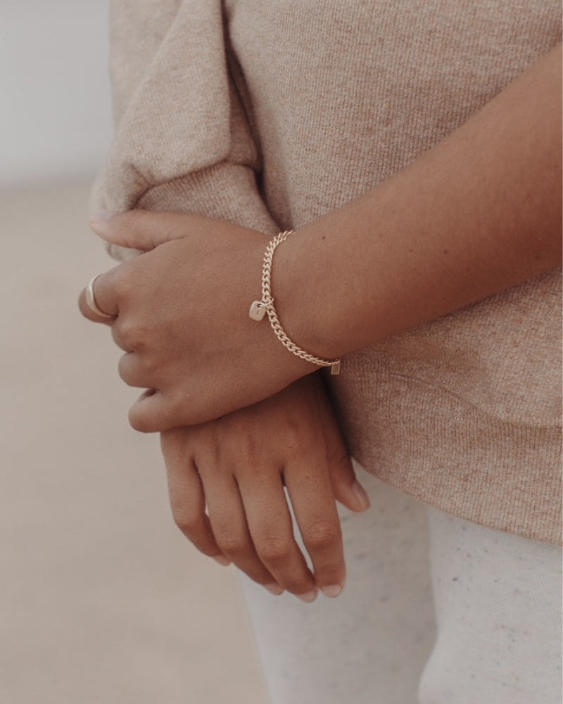 A close-up photo of a woman’s wrist at the beach featuring a Cuban Link Chain with a minimalist rectangular pendant with matching signet ring, styled in fine jewellery fashion by Bluebird Jewellery.