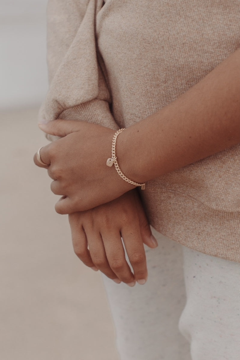 A close-up photo of a woman’s wrist at the beach featuring a Cuban Link Chain with a minimalist rectangular pendant with matching signet ring, styled in fine jewellery fashion by Bluebird Jewellery.